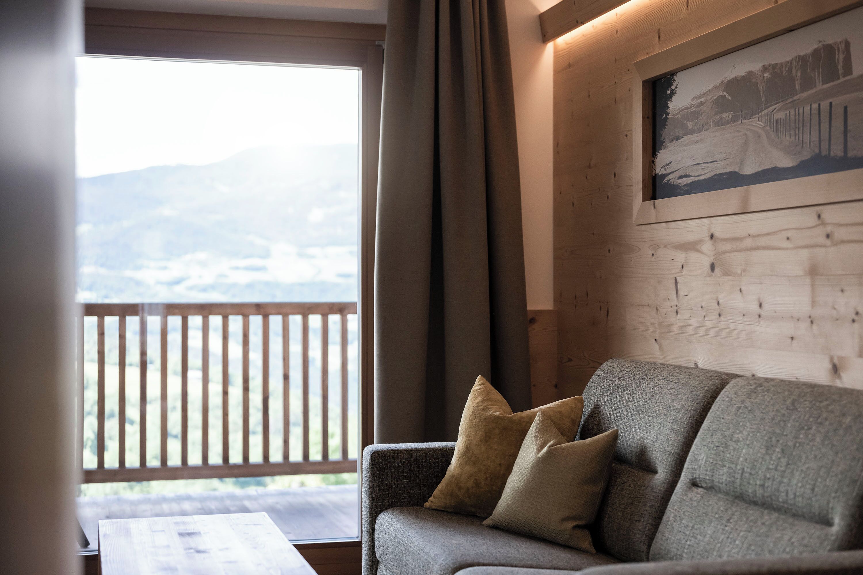 Ein gemütliches Zimmer mit grauem Sofa, einem Holztisch und einem großen Fenster mit Blick auf eine gebirgige Landschaft.