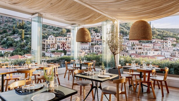 A restaurant with tables set for dining, large windows offering a view of a hillside town.
