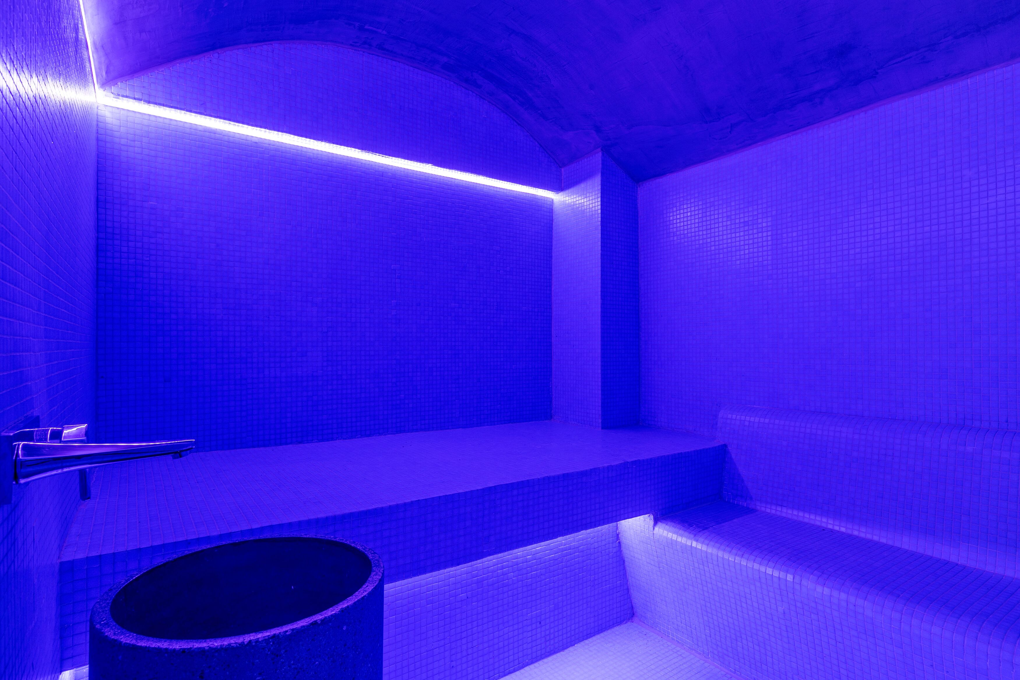 Un baño con tonalidades azules, paredes con azulejos, un banco y un lavamanos.