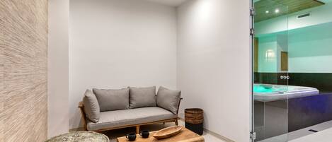 A modern, minimalist bathroom with a glass-enclosed shower, a wooden bench, and a grey sofa.