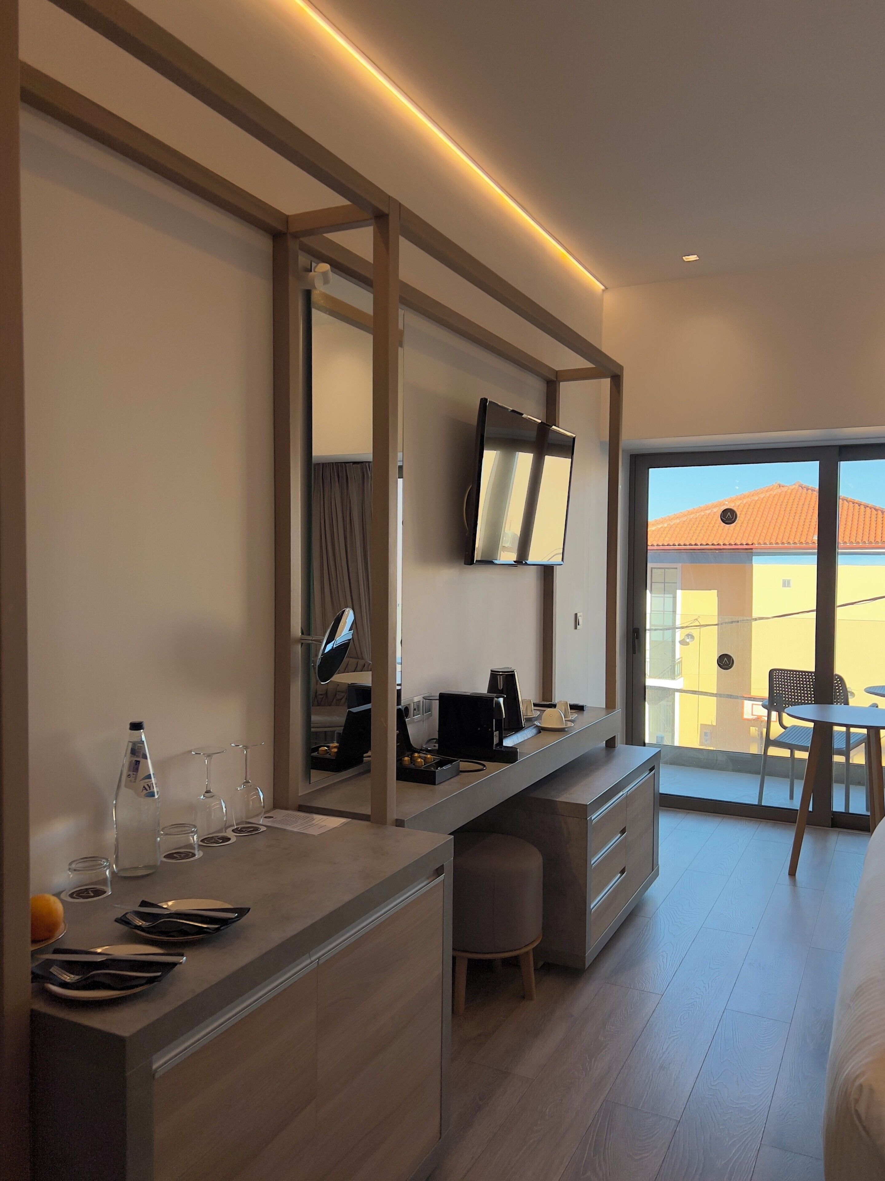 A modern hotel room with a large desk, a flat-screen TV, a dining area with a table and chairs, and a balcony with a view.