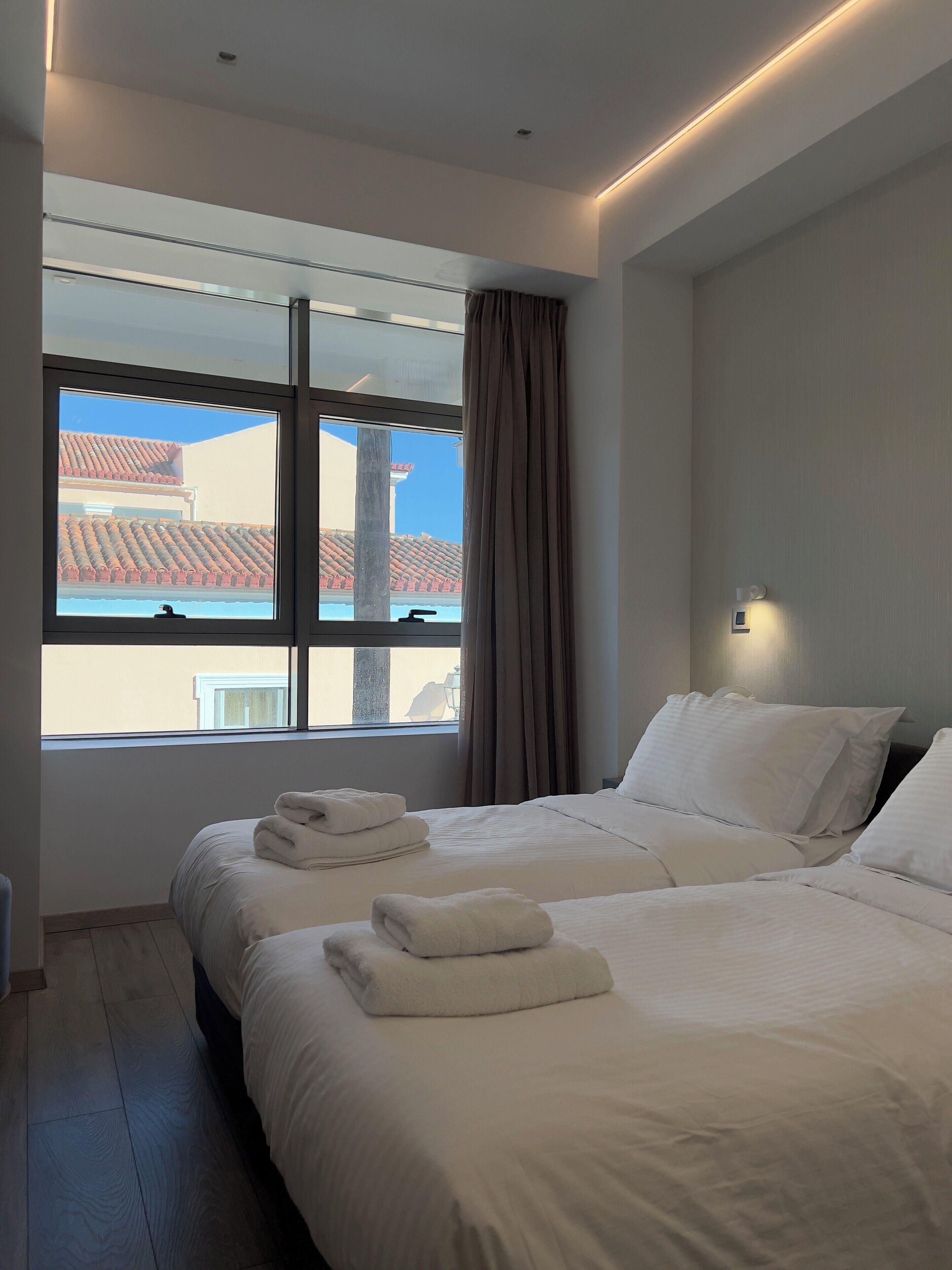 Een hotelkamer met twee bedden, een groot raam met gordijnen, uitzicht op gebouwen en een wandlamp.