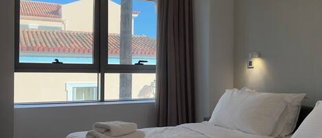 A hotel room with two beds, a large window with curtains, a view of buildings outside, and a wall-mounted light fixture.