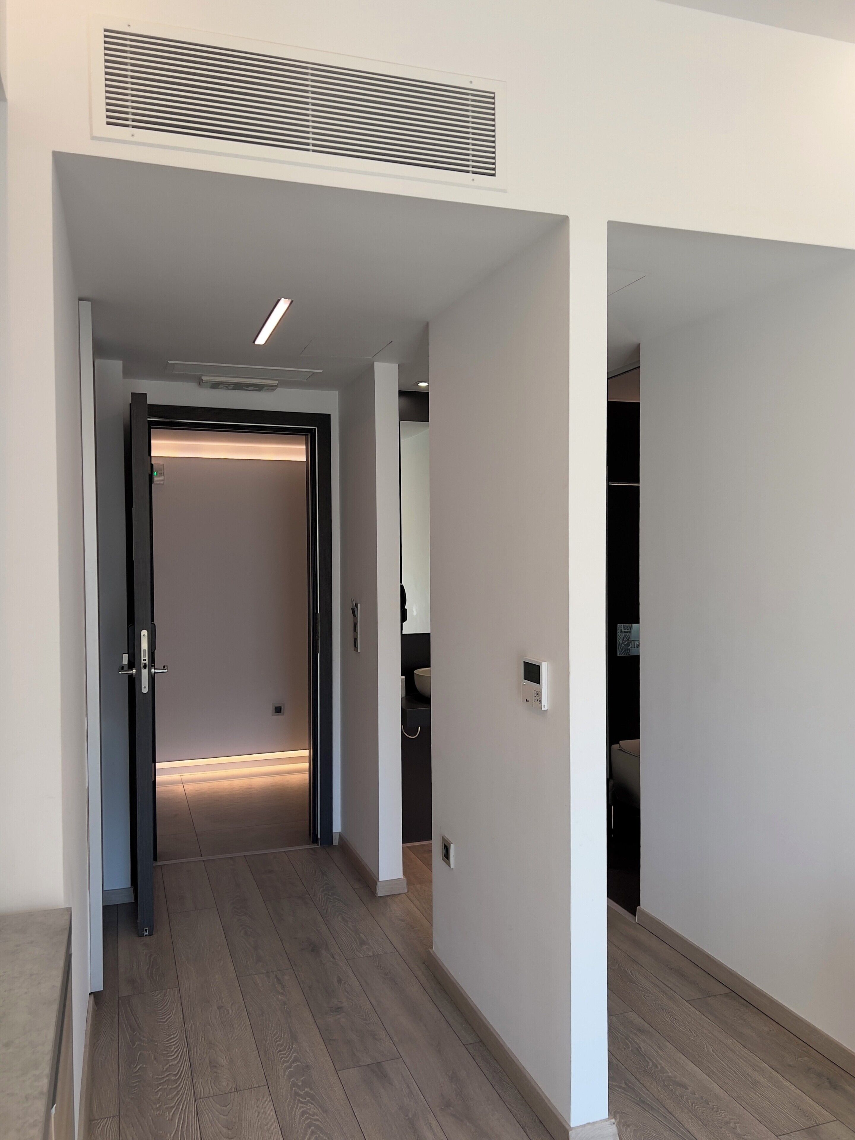 A hallway with a wooden floor, a ceiling vent, and a door leading to a room with a light fixture.