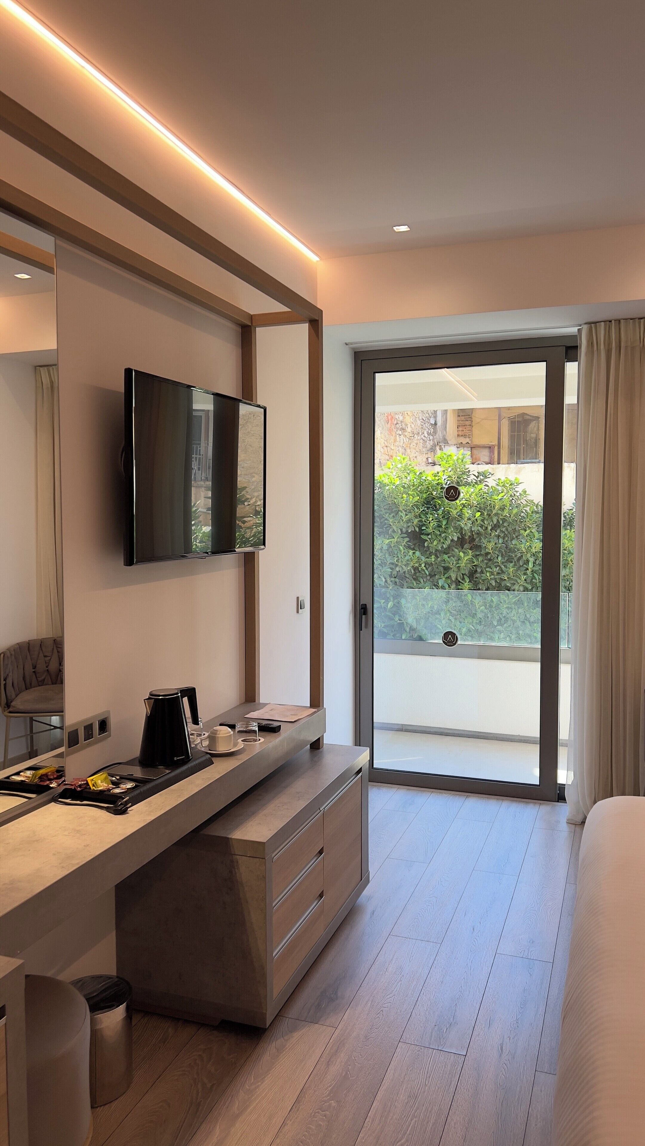 Una habitación de hotel moderna con televisión de pantalla plana, un escritorio integrado con cafetera, un amplio ventanal corredero a un balcón y una cama.