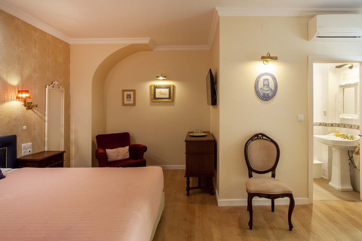 Ein Hotelzimmer mit Bett, Sessel, kleinem Tisch, Wand-Fernseher und einem Badezimmer inklusive Waschbecken und Spiegel.