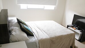 A neatly made bed with a white and beige bedspread, a television on a stand, and a window providing natural light. - Buda Apartments (San Rafael)