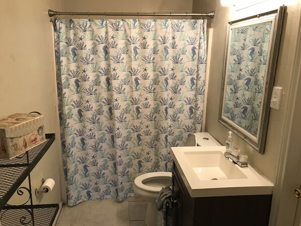 Baño con cortina de ducha estampada con motivos de caballitos de mar y coral, inodoro, lavabo con espejo y repisa con artículos de aseo.