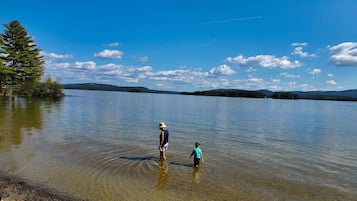 A person and a child standing in shallow water by a lake with trees and hills in the background.