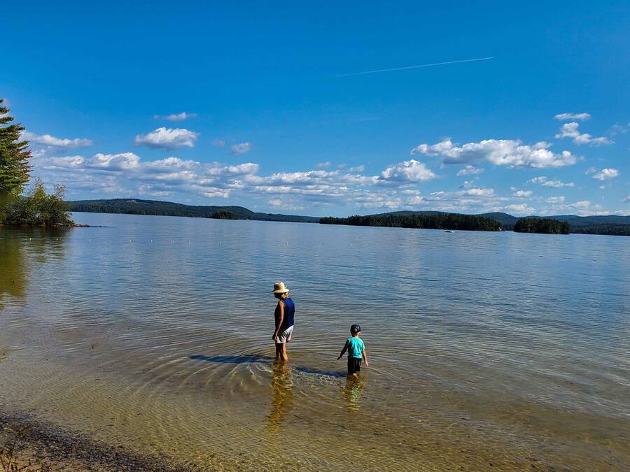 A person and a child standing in shallow water by a lake with trees and hills in the background.