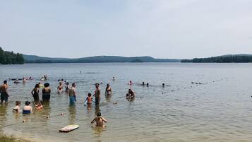 A lakeside scene with people swimming and standing in the water, kayaks, and a sandy shore.