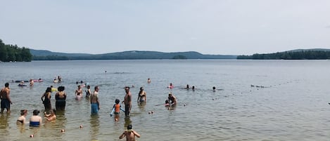 Une scène au bord d’un lac, avec des personnes qui nagent et qui se tiennent dans l’eau, des kayaks et une plage de sable.