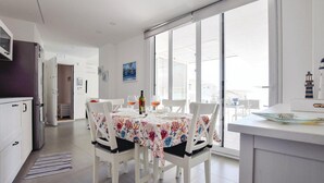 A dining area with a table set for four, white chairs, and a view through large windows. - Lovely apartment in Marina di Ragusa (Marina di Ragusa)
