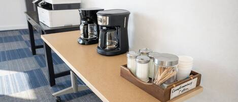 A coffee station with a coffee maker, a grinder, and a container labeled 'Complimentary Coffee.'