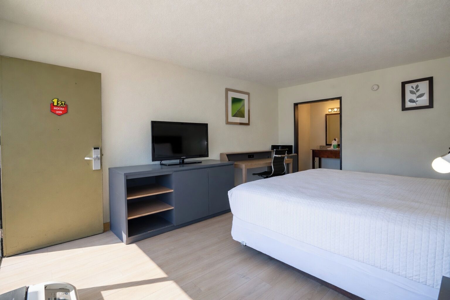 A hotel room with a bed, a TV on a cabinet, a desk with a chair, a mirror, and a door with a sign.