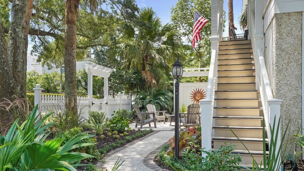 A garden pathway leading to a white gazebo with outdoor furniture, surrounded by lush greenery and a palm tree.