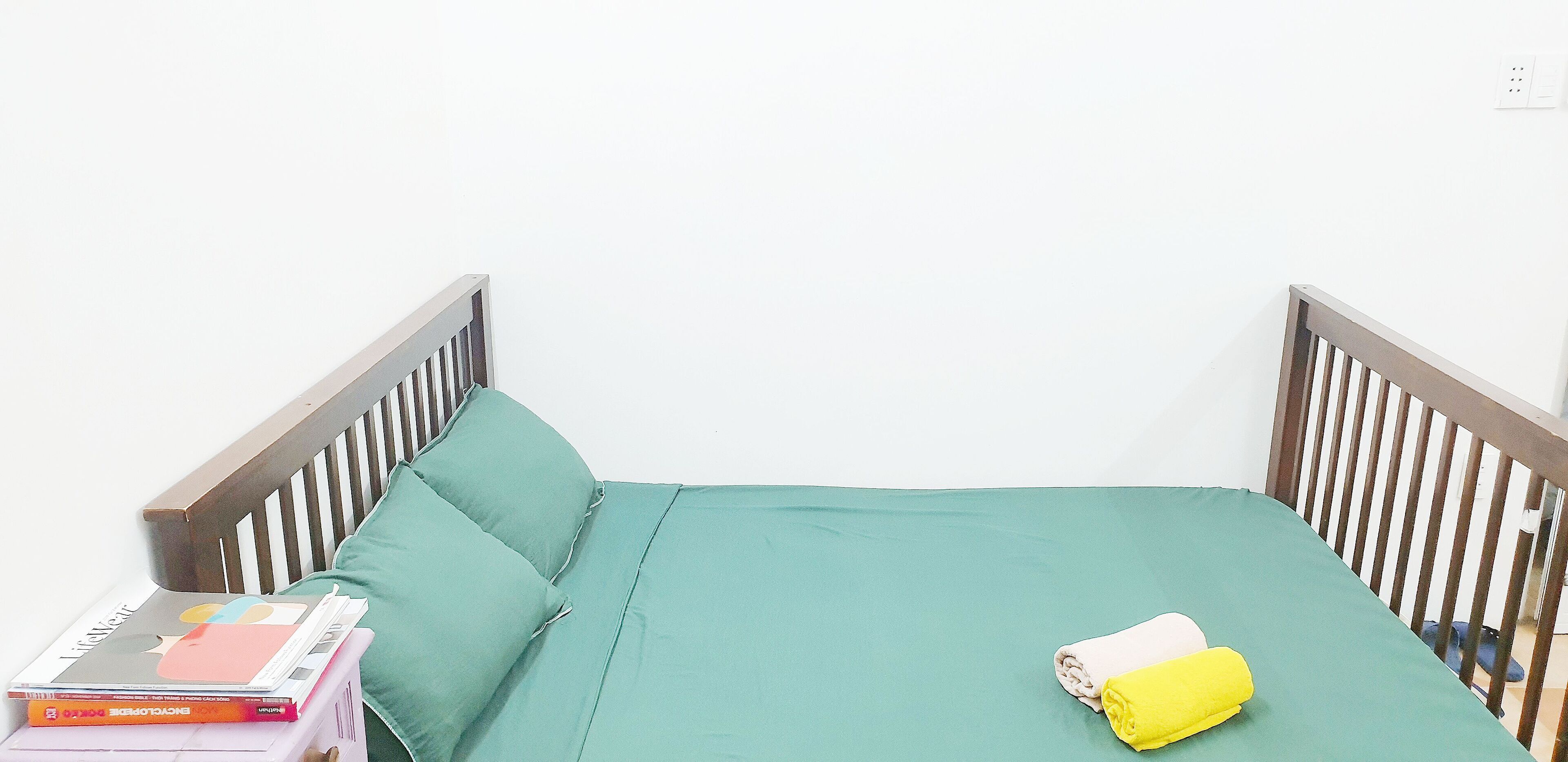 A small bed with a green mattress, a wooden headboard, and a pillow. There are books stacked on the side and a yellow towel on the bed.