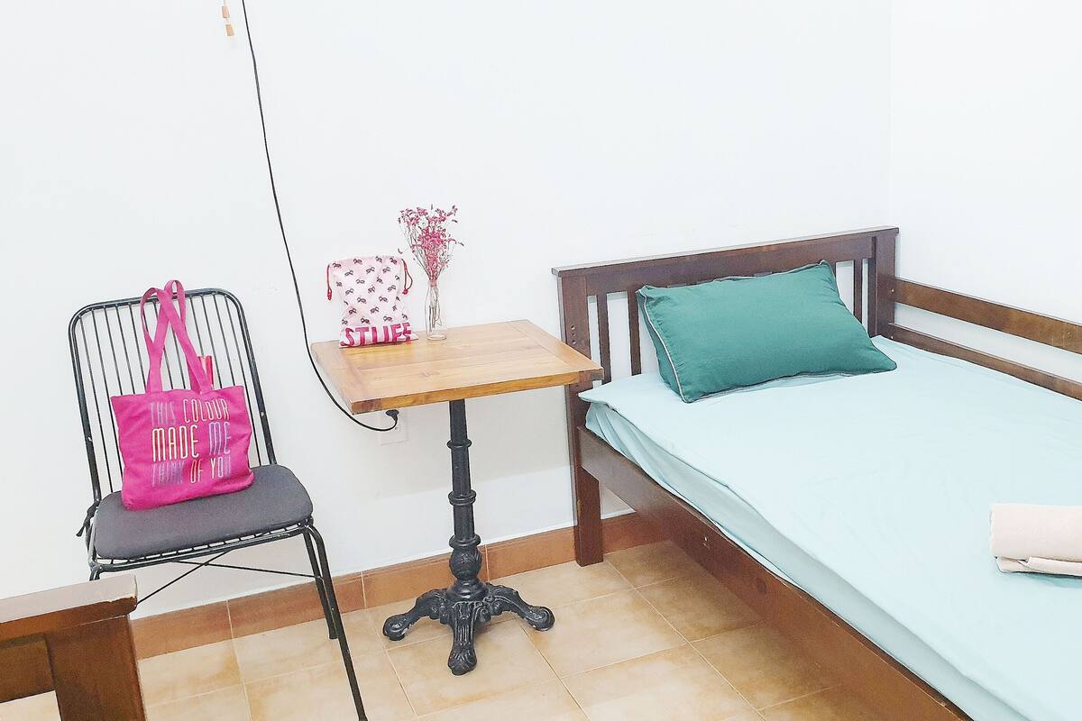 Una cama individual con estructura de madera, una mesita de madera con una silla, un bolso rosa y un florero con flores secas.