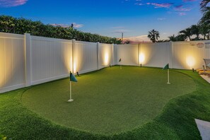 A well-lit backyard with artificial turf and two golf flags, surrounded by a white fence.