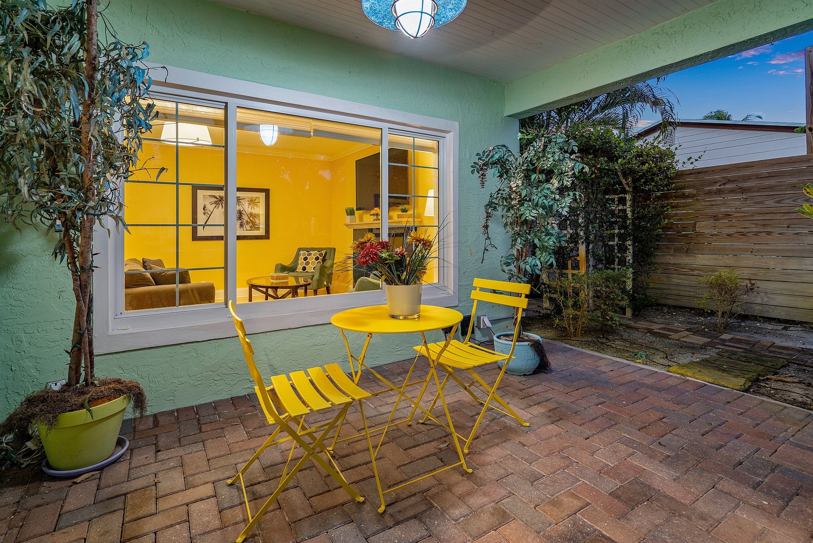 Un patio con sillas amarillas y una mesa, una planta en maceta, y una ventana con vista al interior.