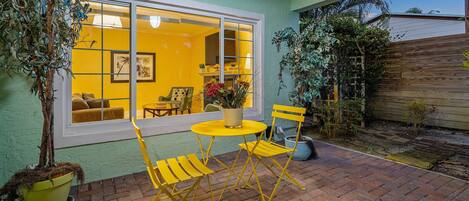 Un patio con sillas amarillas y una mesa, una planta en maceta, y una ventana con vista al interior.