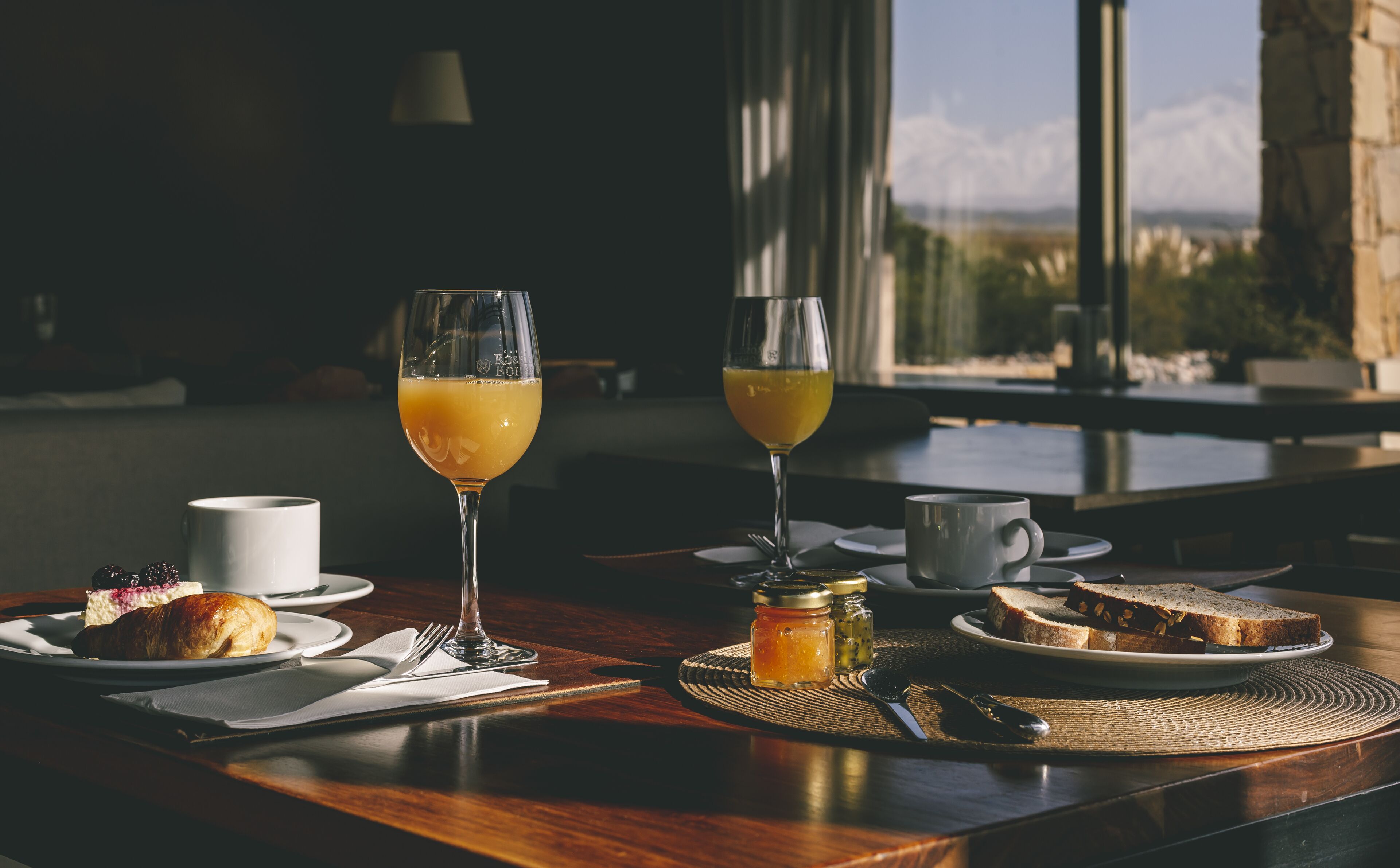 Une table en bois dressée pour le petit-déjeuner, avec des croissants, des toasts et deux verres de jus d’orange.