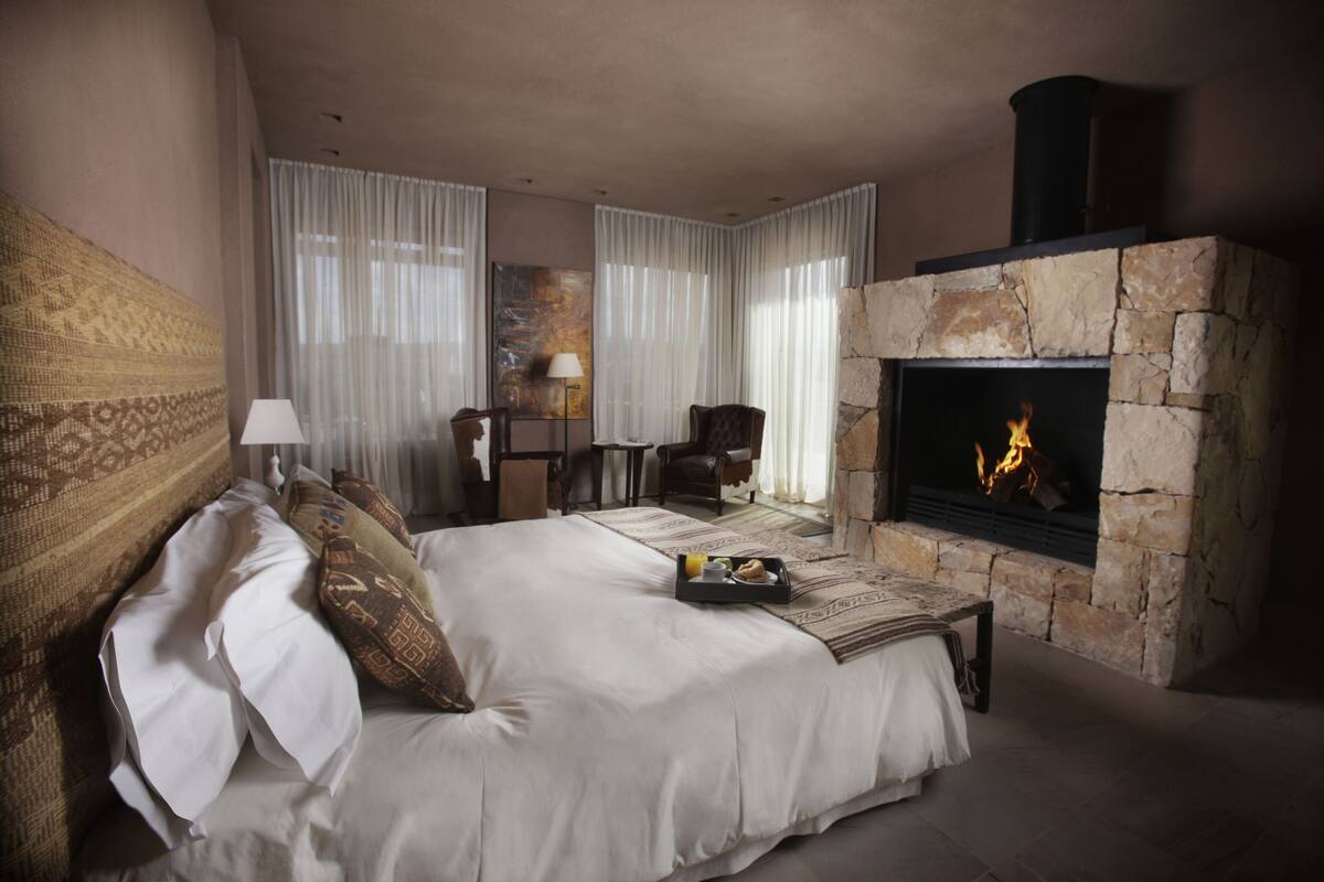 A bedroom with a stone fireplace, a bed with white linens, a side table with a tray of breakfast items, and a chair.