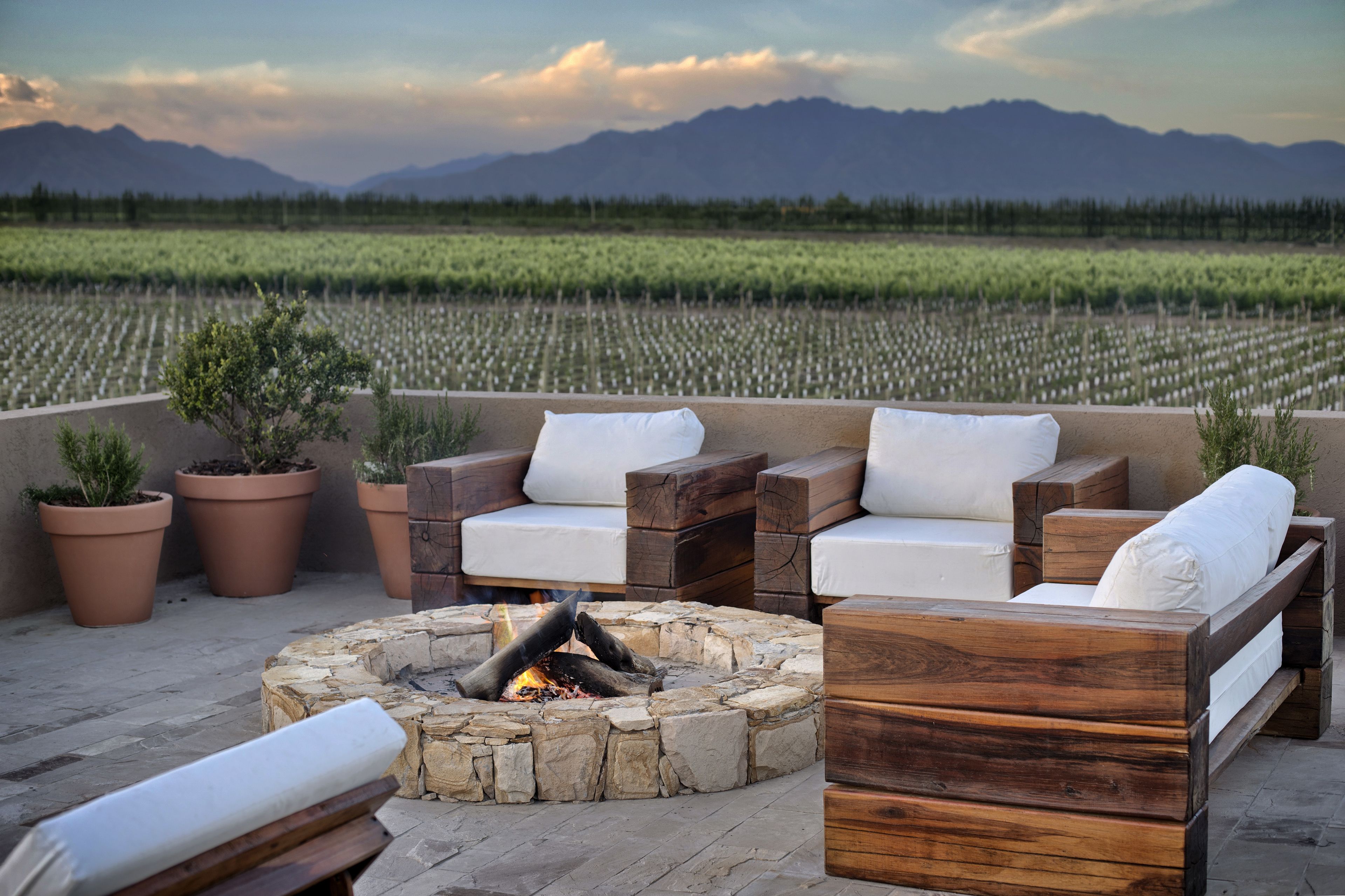 An outdoor seating area with a stone fire pit, wooden furniture, and potted plants.