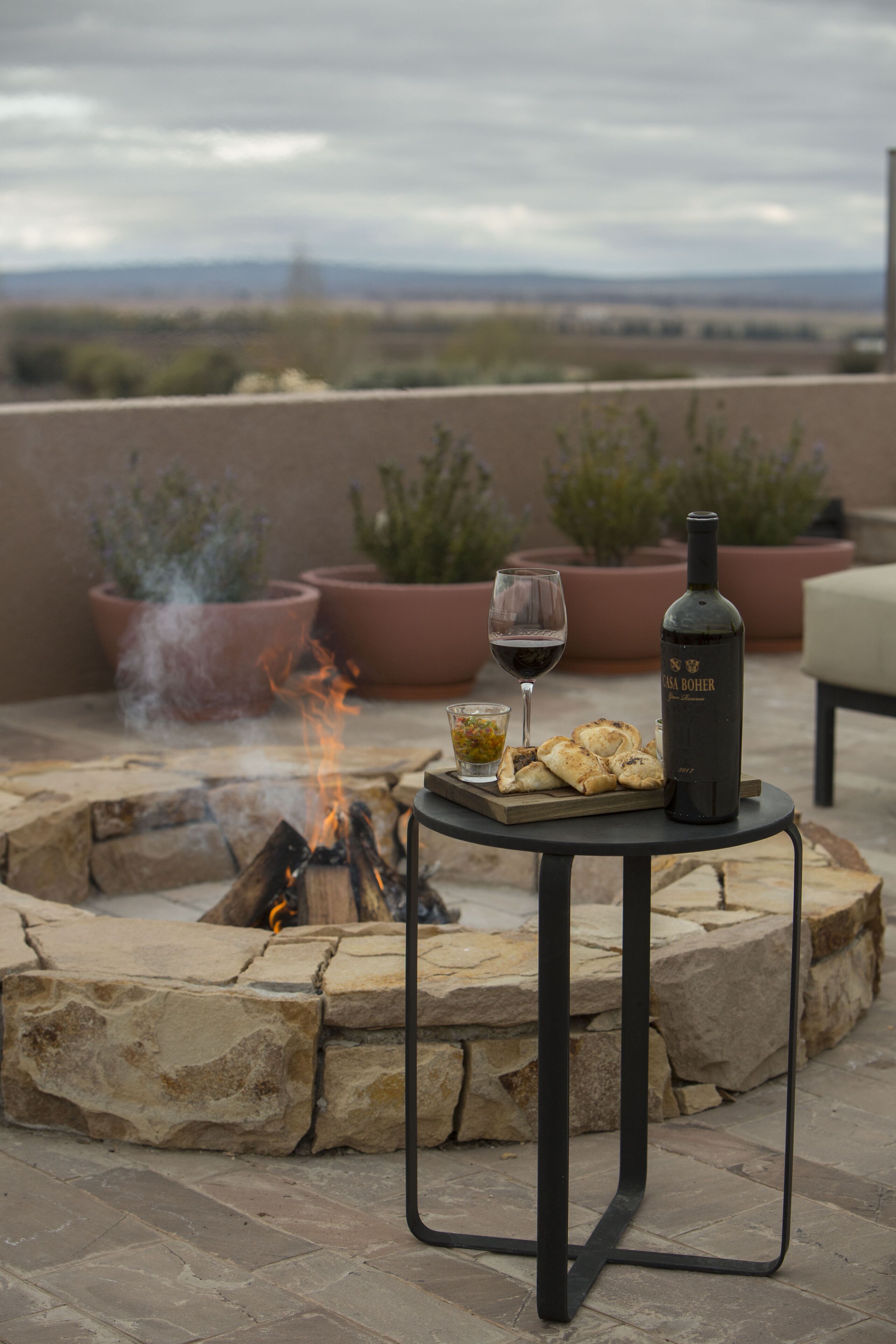 A stone fire pit with a table and chairs, a bottle of wine, and a glass of wine on a patio.