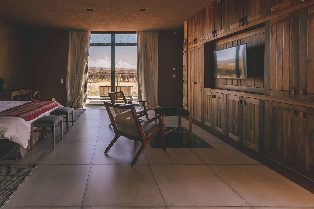 A hotel room with a large bed, a TV mounted on the wall, a dining area with chairs, and a view of a mountainous landscape through the window.