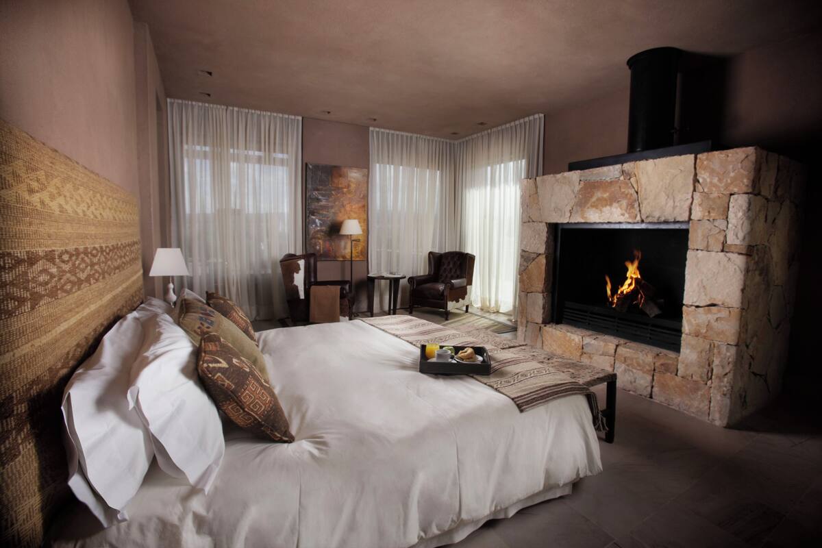 A bedroom with a stone fireplace, a bed with white linens, a side table with a tray of breakfast items, and a chair.