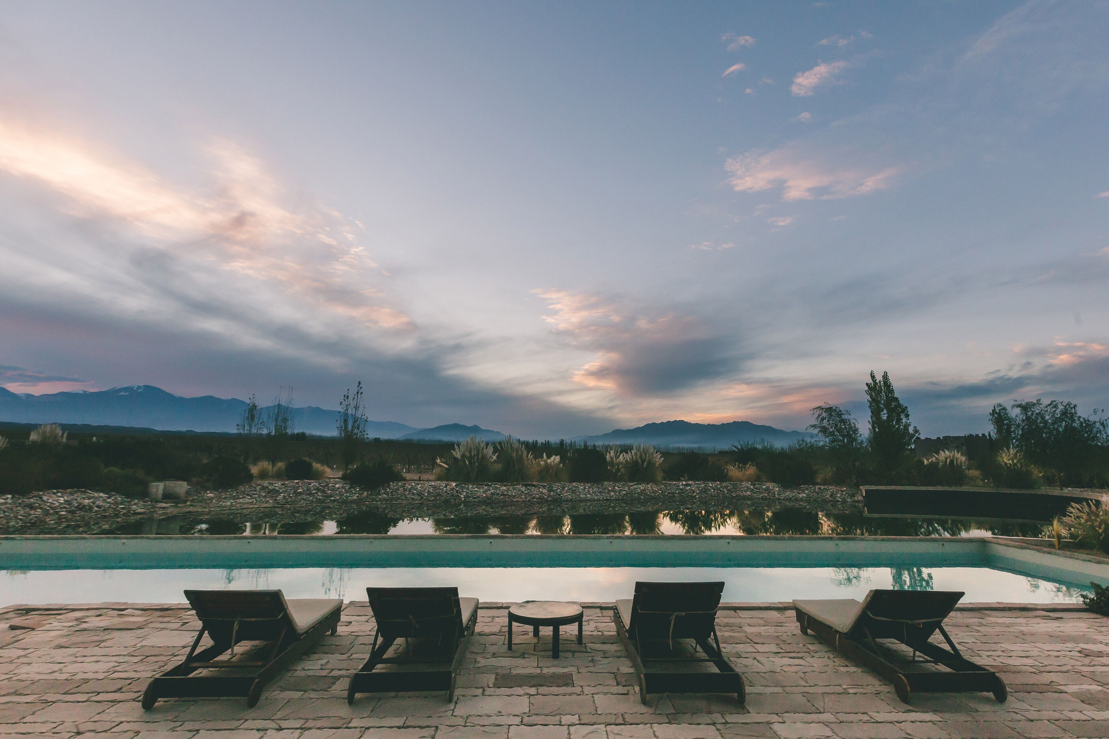 A poolside area with lounge chairs and a table, overlooking a calm pool and distant mountains.