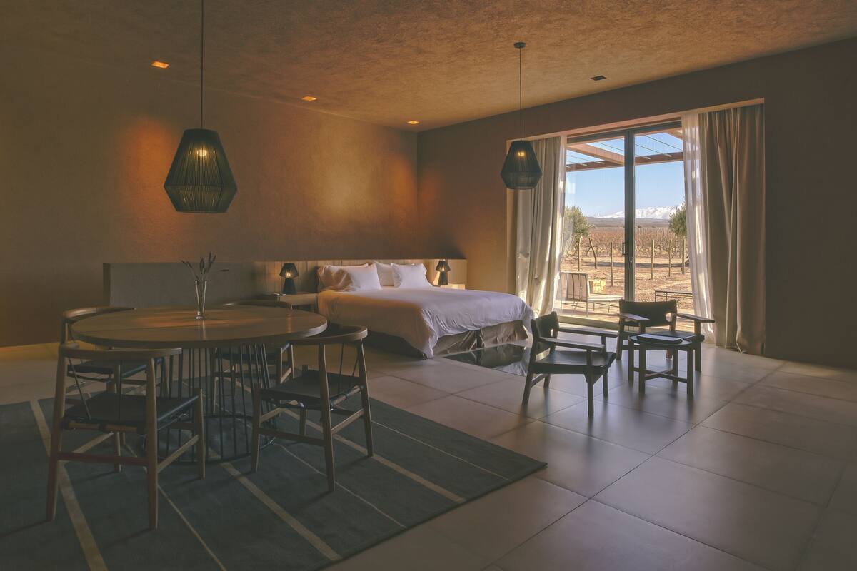 A modern hotel room with a bed, a dining area with a round table and chairs, and a view of the outdoors through a large window.