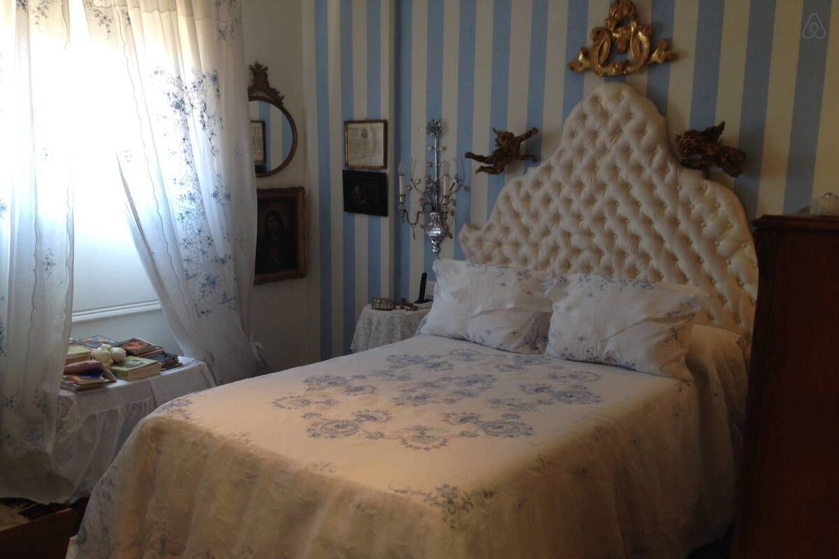 Quarto com cama, mesa de cabeceira, janela com cortinas e papel de parede listrado com decorações.
