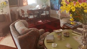 Una sala de estar con un sofá estampado, una mesa de comedor con sillas, un jarrón con flores amarillas y un cuadro en la pared.