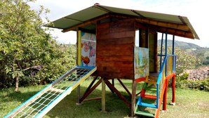 A wooden playhouse with a slide and stairs, surrounded by greenery.