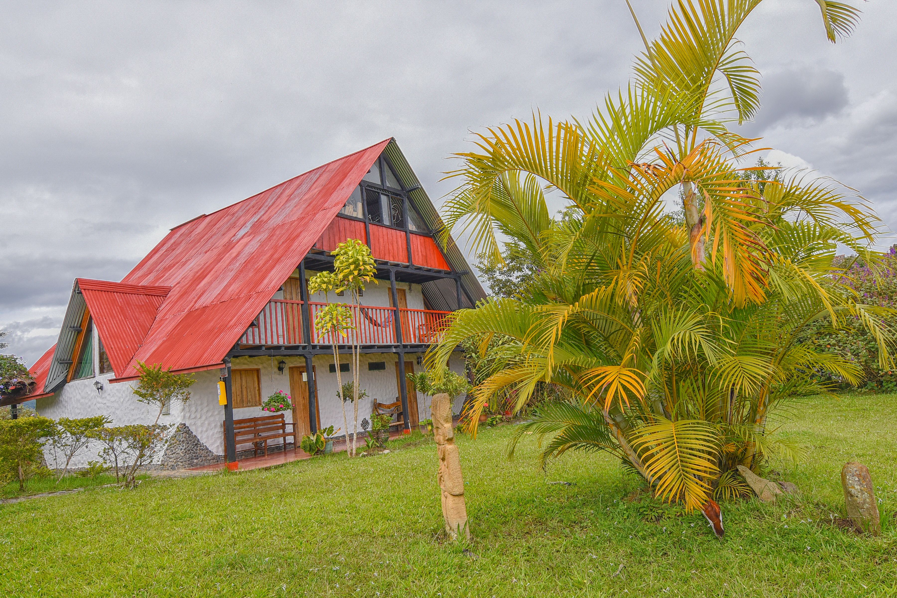 A house with a sloped red roof, a balcony, and a garden with palm trees.