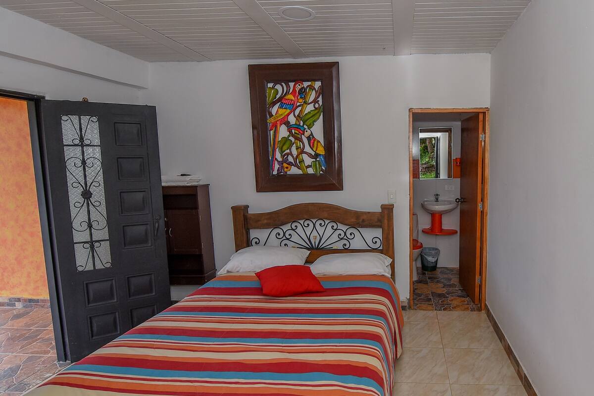 A bedroom with a bed, a wooden headboard, a colorful striped bedspread, a red pillow, a door with a decorative glass panel, a wall-mounted painting, and a bathroom visible through an open door.