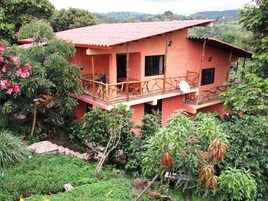Una casa de dos pisos con techo rojo, rodeada de exuberante follaje y árboles.