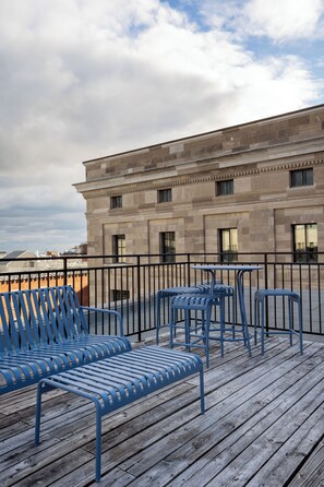 A rooftop terrace with blue metal chairs and tables, a stone building in the background, and a cloudy sky.