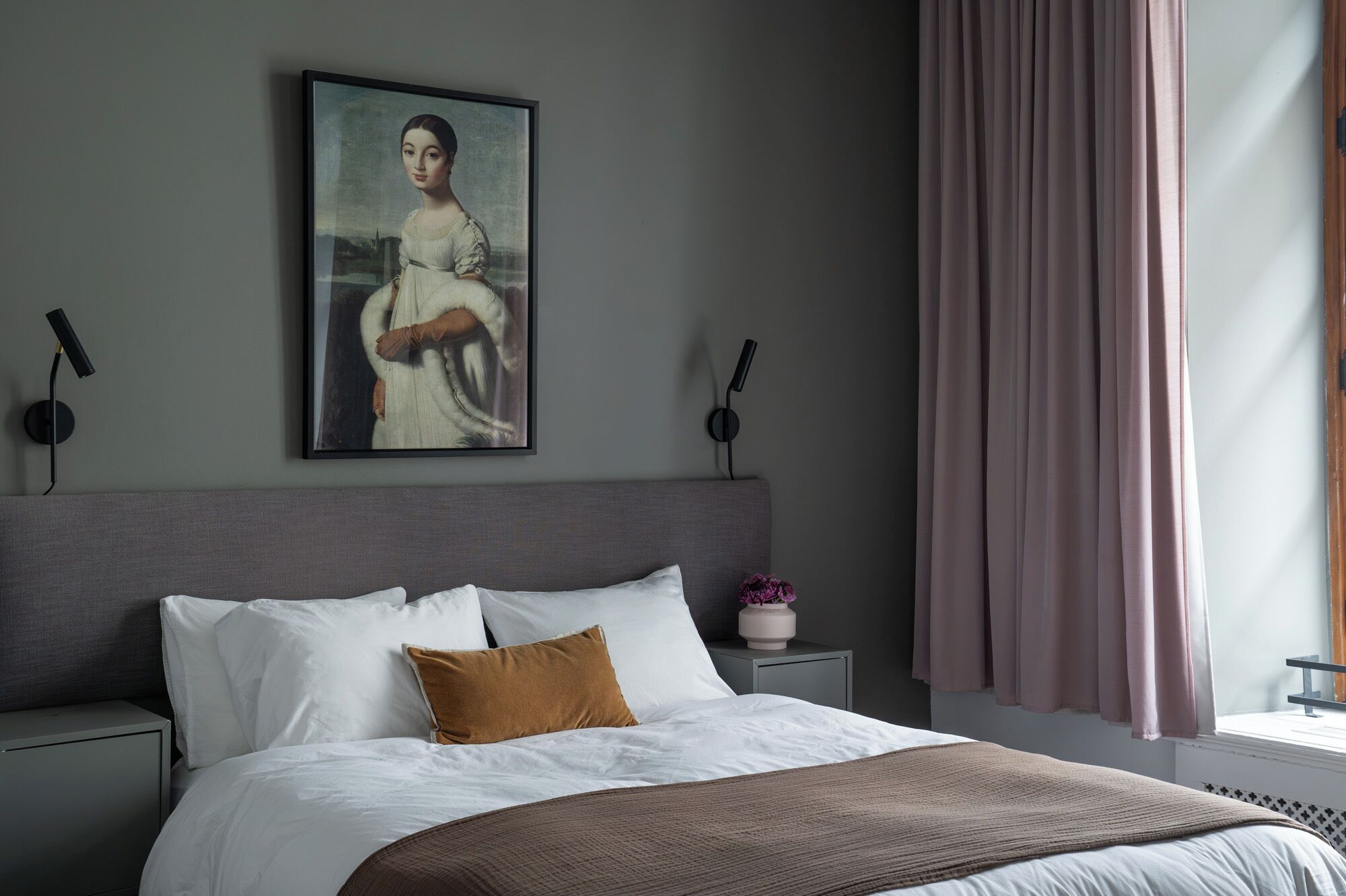 Un dormitorio con cama, mesita de noche, un cuadro y ventana con cortinas.
