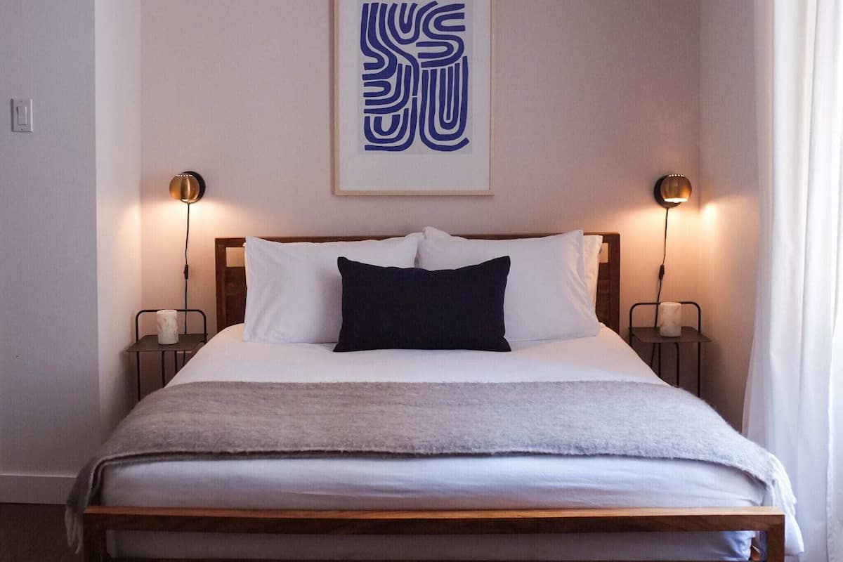 A neatly made bed with a dark pillow, two bedside lamps, and a framed abstract artwork on the wall.