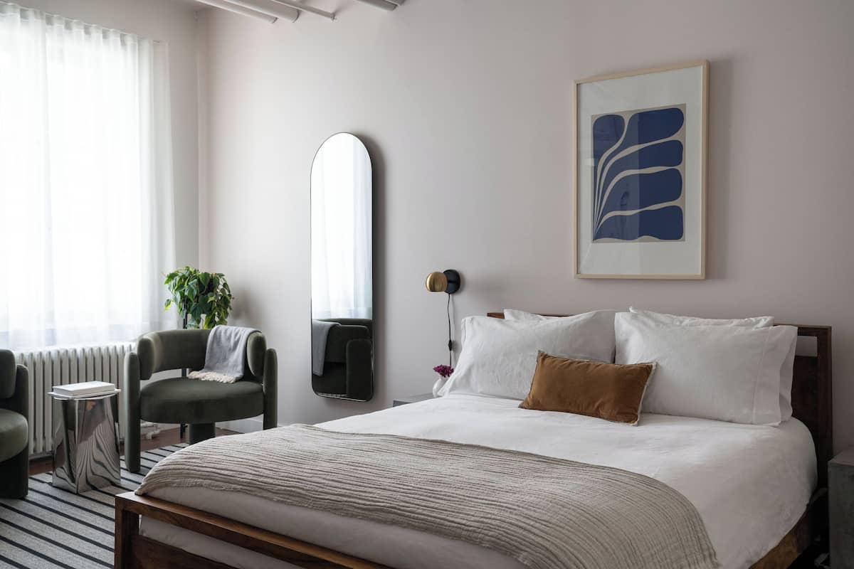 A hotel room with a large bed, a bedside table, a chair, a mirror, and a framed abstract artwork on the wall.