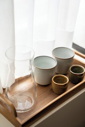 Una bandeja con cuatro tazas de cerámica y una jarra de vidrio sobre una superficie de madera, cerca de una ventana con cortinas translúcidas.