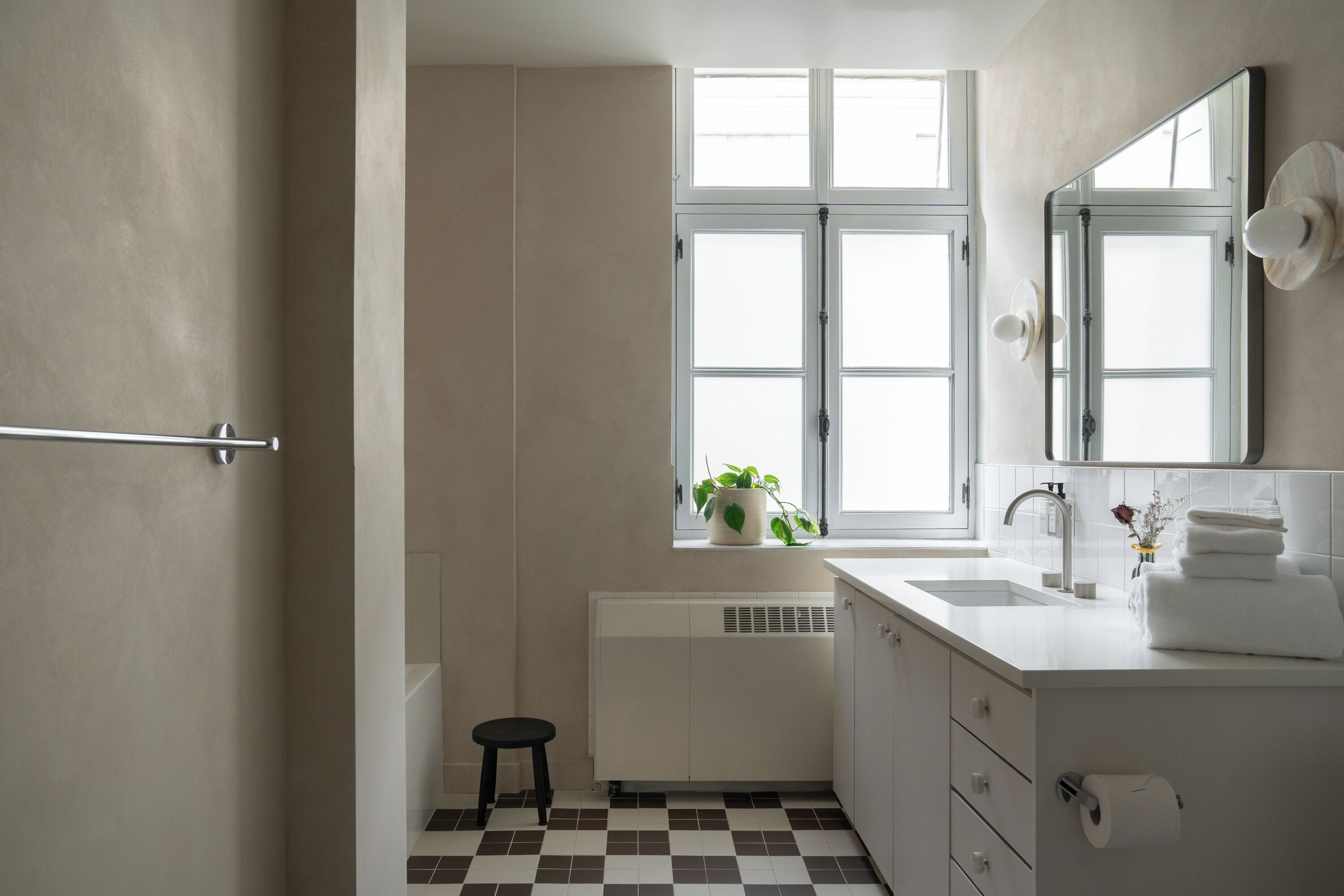 Un bagno con un lavandino bianco, uno specchio, una finestra con stipiti bianchi, un termosifone e uno sgabello nero.