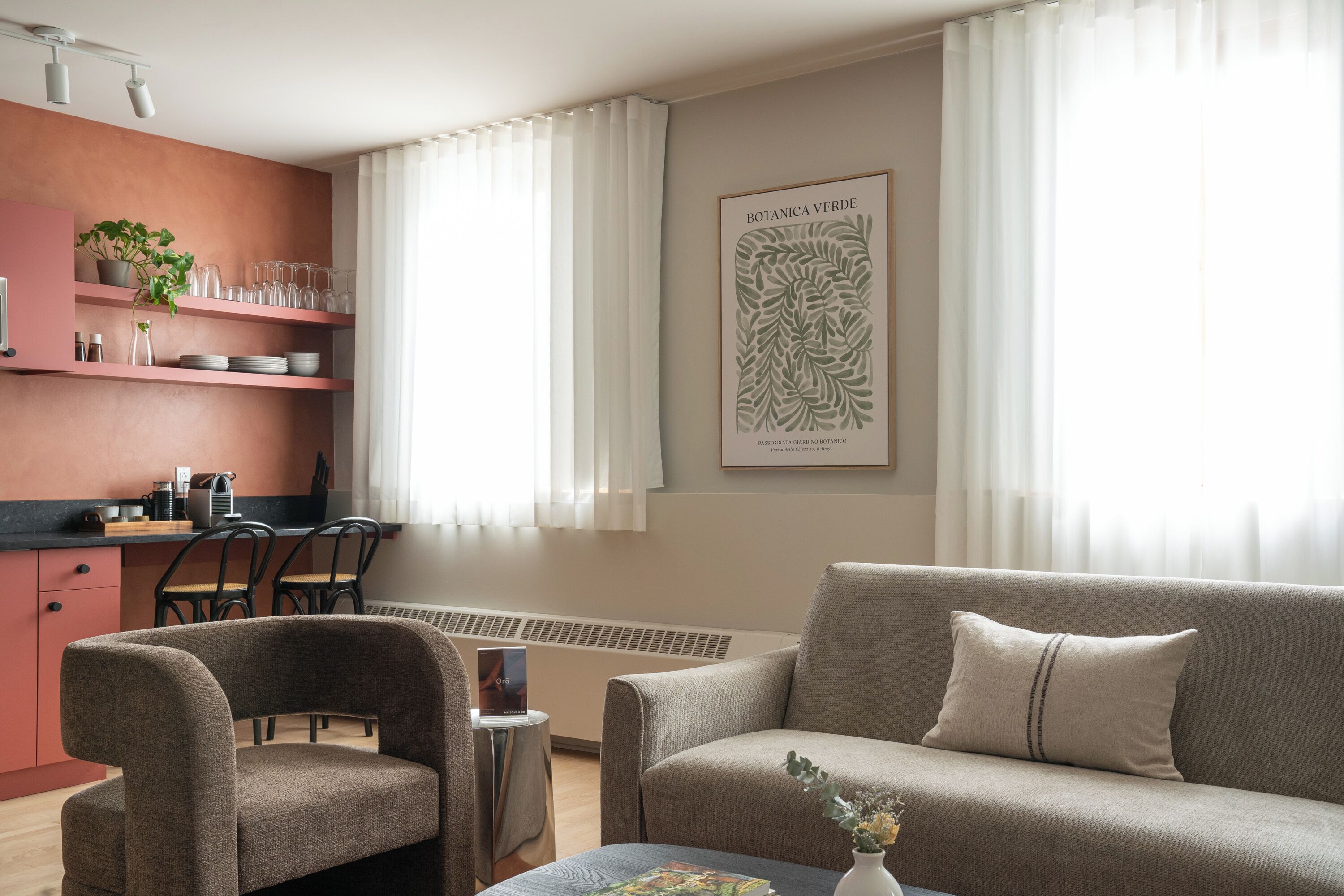Ein modernes Wohnzimmer mit einer Couch, Sesseln, einem Couchtisch, einer Küchenzeile mit Arbeitsplatte und Regalen sowie einem großen botanischen Wandbild.