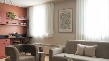Ein modernes Wohnzimmer mit einer Couch, Sesseln, einem Couchtisch, einer Küchenzeile mit Arbeitsplatte und Regalen sowie einem großen botanischen Wandbild.