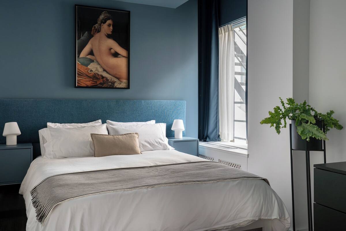 A bedroom with a large bed, a headboard, a nightstand, a bedside table, a lamp, a painting, a window with curtains, and a potted plant.