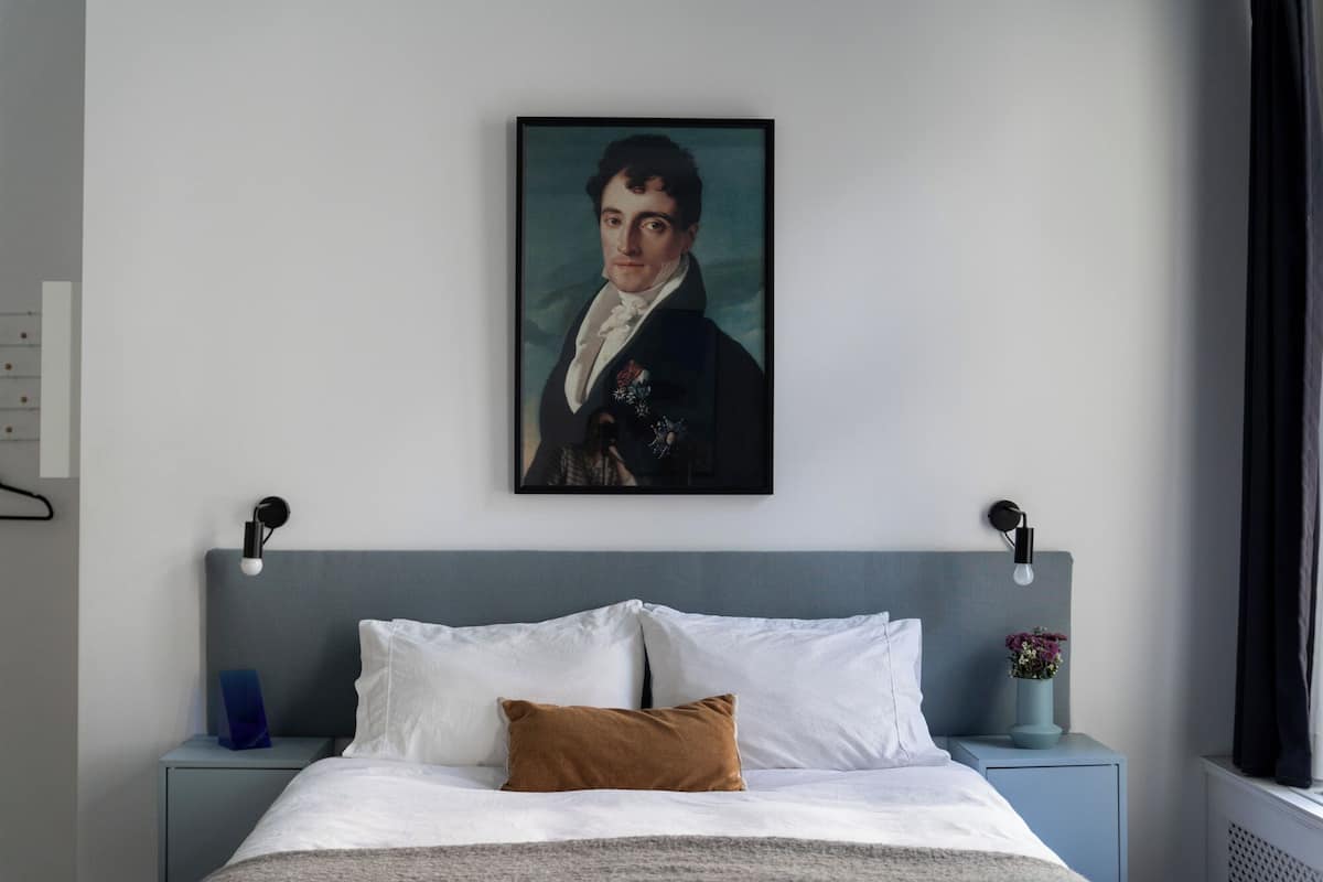 A bedroom with a bed, a nightstand, a wall-mounted painting, and a bedside table with a vase.