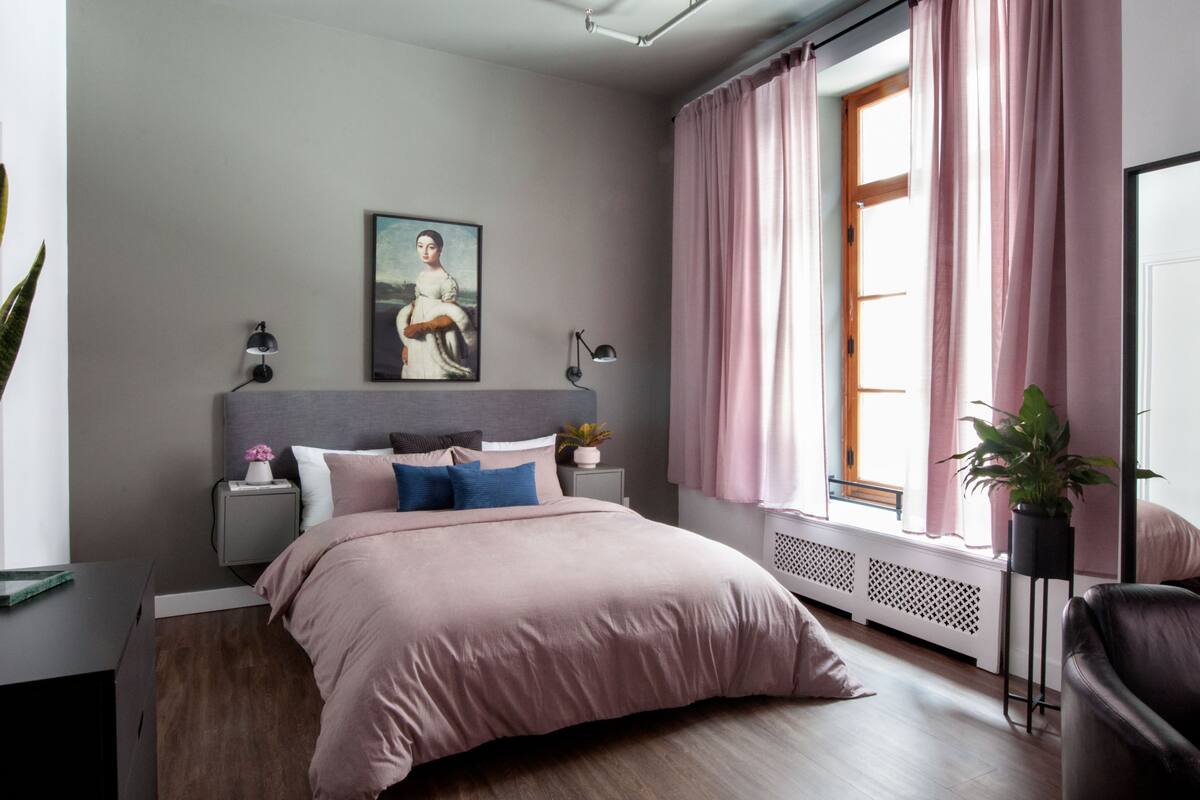 Un dormitorio con cama, mesitas de noche, una planta y un cuadro en la pared.